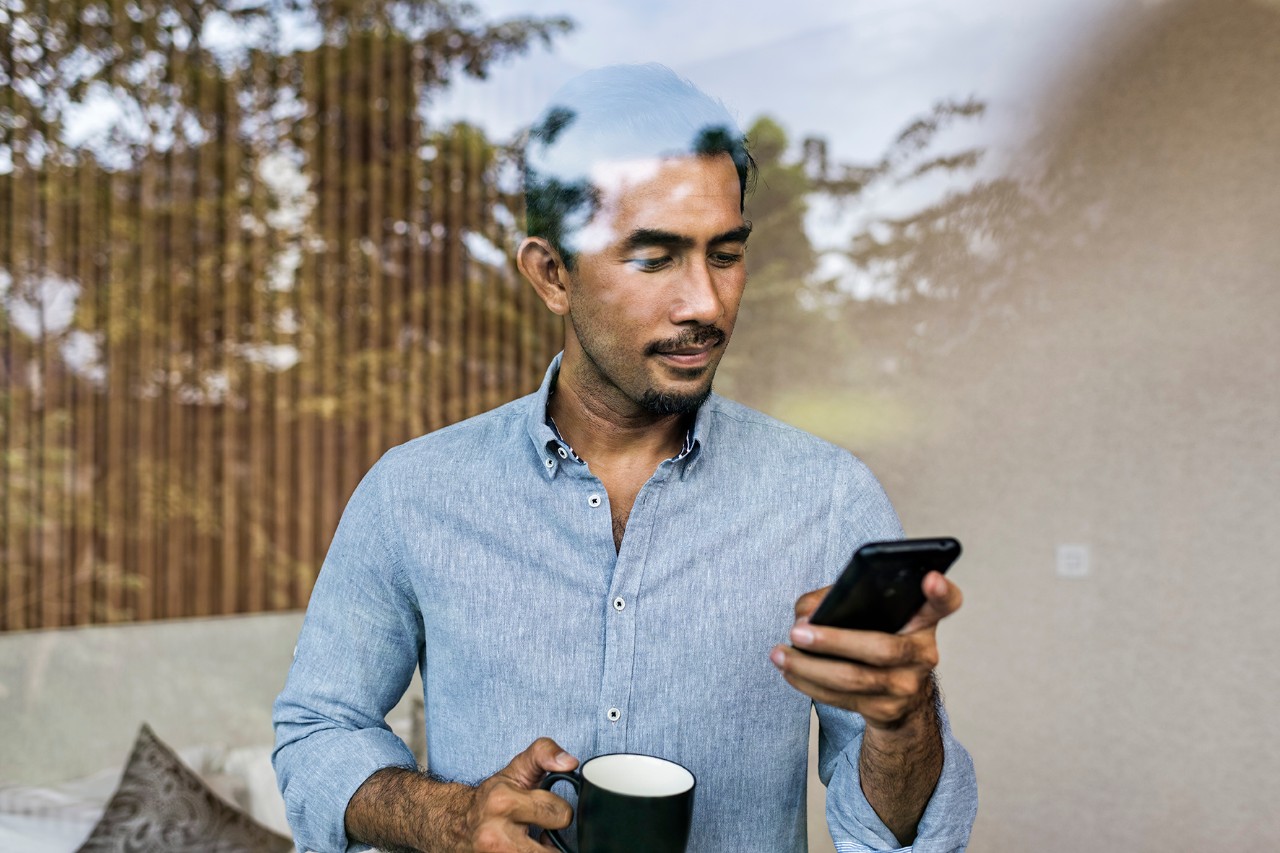 Asian man holding iphone Asia, Indonesia, Jakarta, Asian businessman working from home, using smartphone and drinking coffee, while standing at bedroom window of modern house