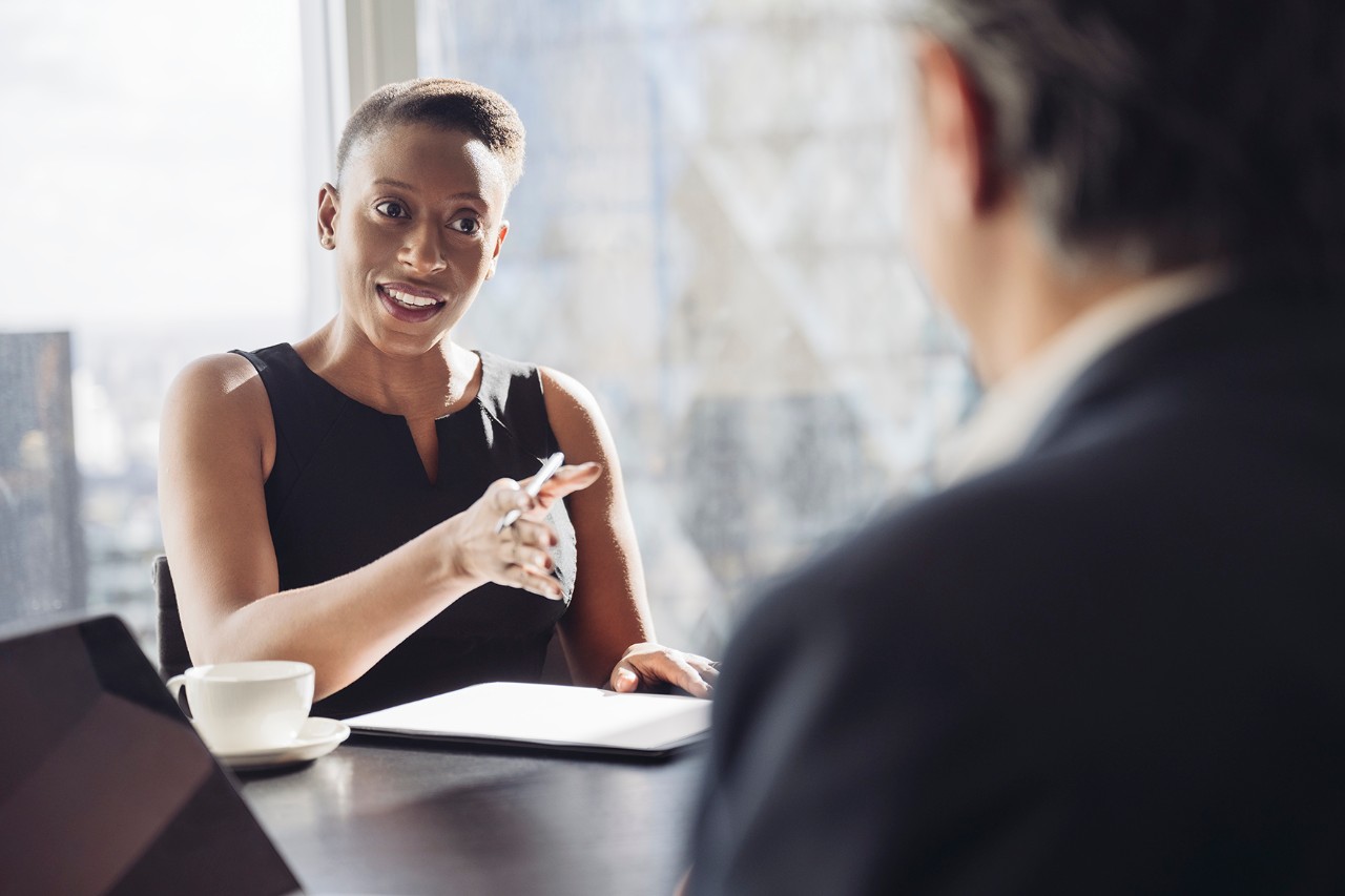 Woman interview Over the shoulder view of well-dressed British executive in mid 30s sitting at conference table in sunny meeting room and smiling as she makes her point.